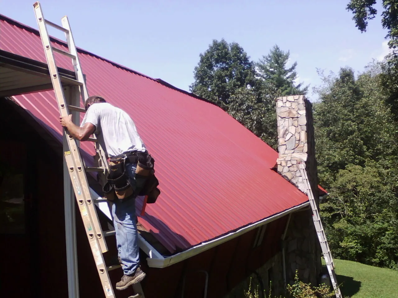 Roofer on a ladder beside a red metal roof and stone chimney — Soffit & Fascia Repair by Roofing Brothers in Hannaford